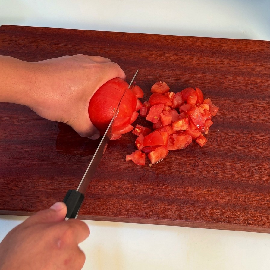 Eine Hand schneidet mit einem Kochmesser eine geschälte rote Tomate auf einem dunklen Holzschneidebrett.