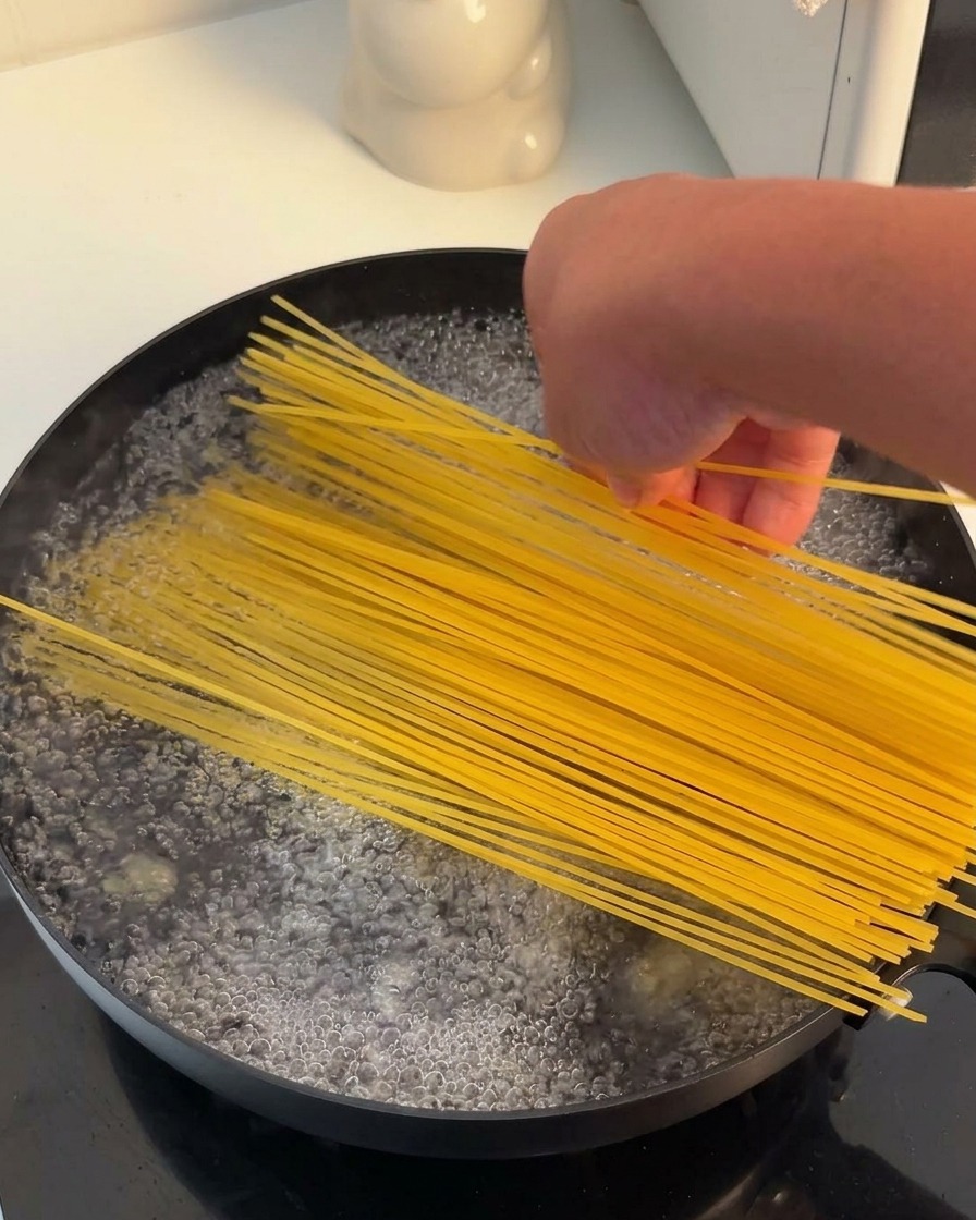 Eine Hand gibt ein Bündel trockener Spaghetti in einen schwarzen Topf mit kochendem Wasser.
