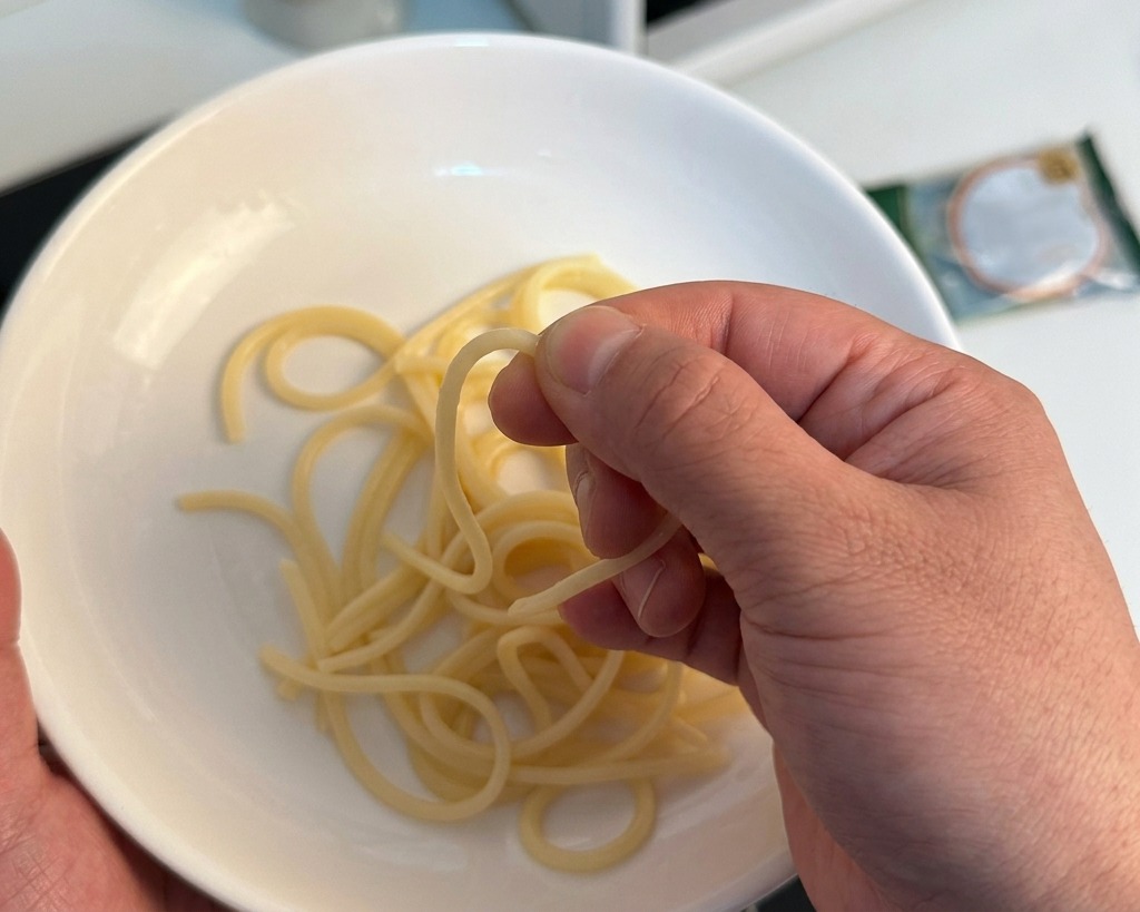 Eine Hand zerdrückt leicht eine einzelne gekochte Spaghetti über einer weißen Schüssel, um die Konsistenz zu prüfen.