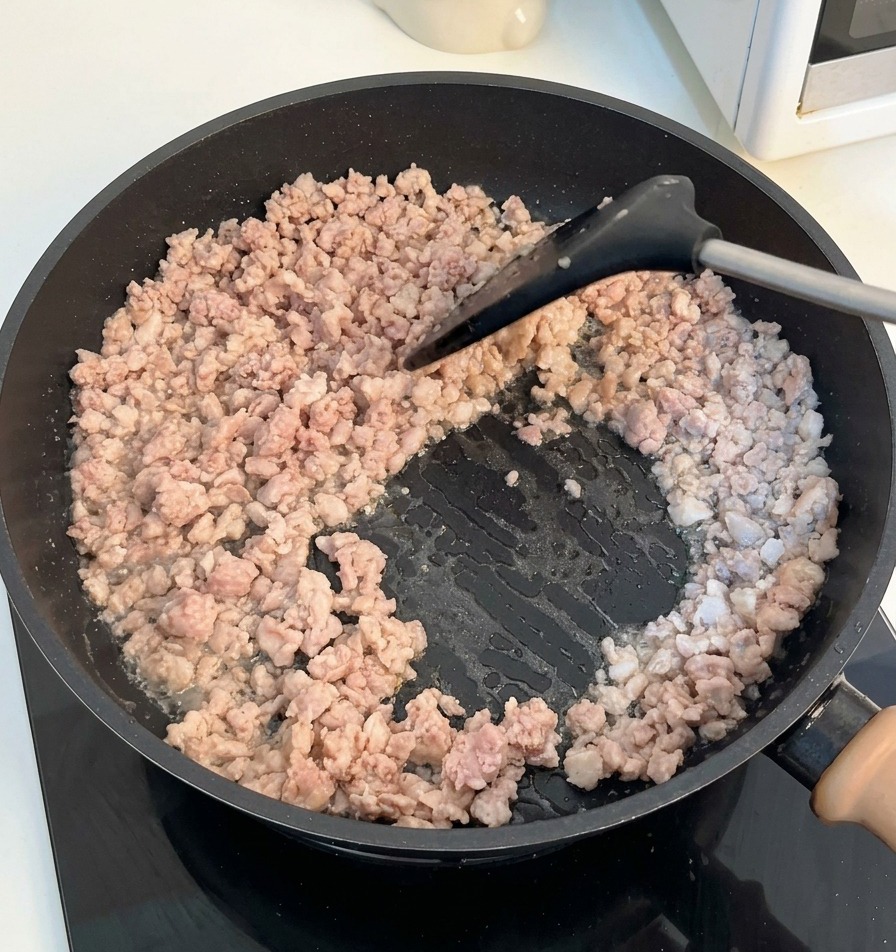 Ein schwarzer Pfannenwender zerkleinert und rührt das gebräunte Hackfleisch in einer dunklen Pfanne.
