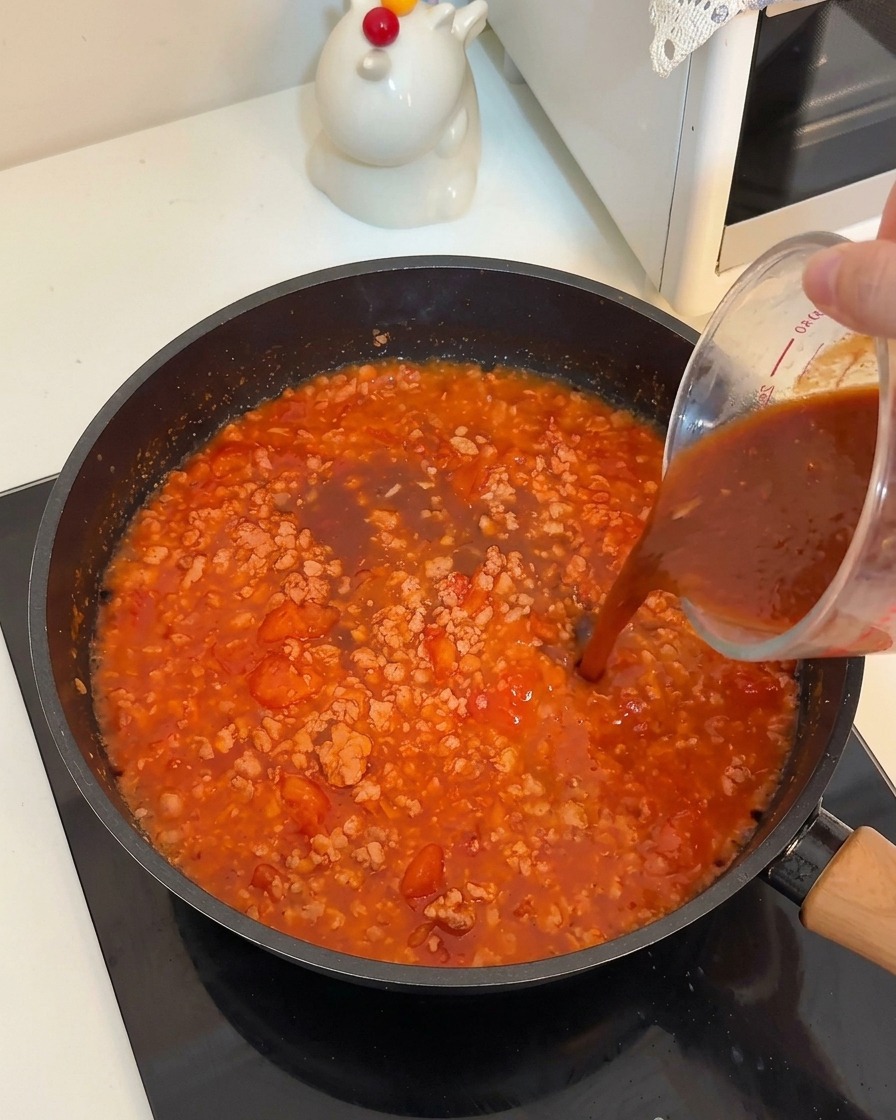 Eine dunkle Würzsoße wird aus einem Messbecher über die köchelnde Fleisch-Tomaten-Mischung gegossen.