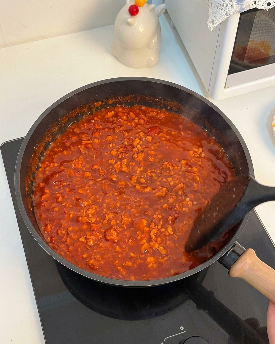 Eine dicke, leuchtend rote Tomaten-Hackfleischsoße blubbert in einer Pfanne und wird umgerührt.