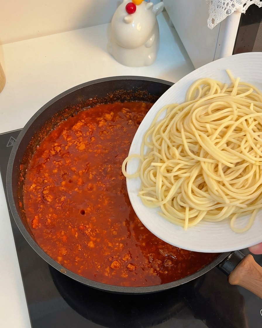 Gekochte Spaghetti gleiten in eine Pfanne mit sämiger Tomaten-Hackfleischsoße.