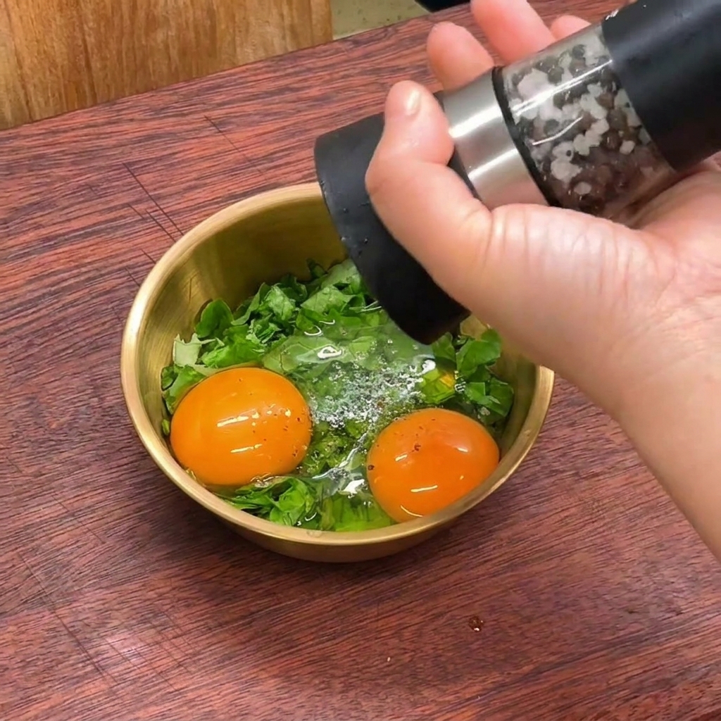 Eine Hand benutzt eine Pfeffermühle, um frischen schwarzen Pfeffer in eine kleine goldene Schüssel mit rohen Eiern und geschnittenem grünen Salat zu mahlen.