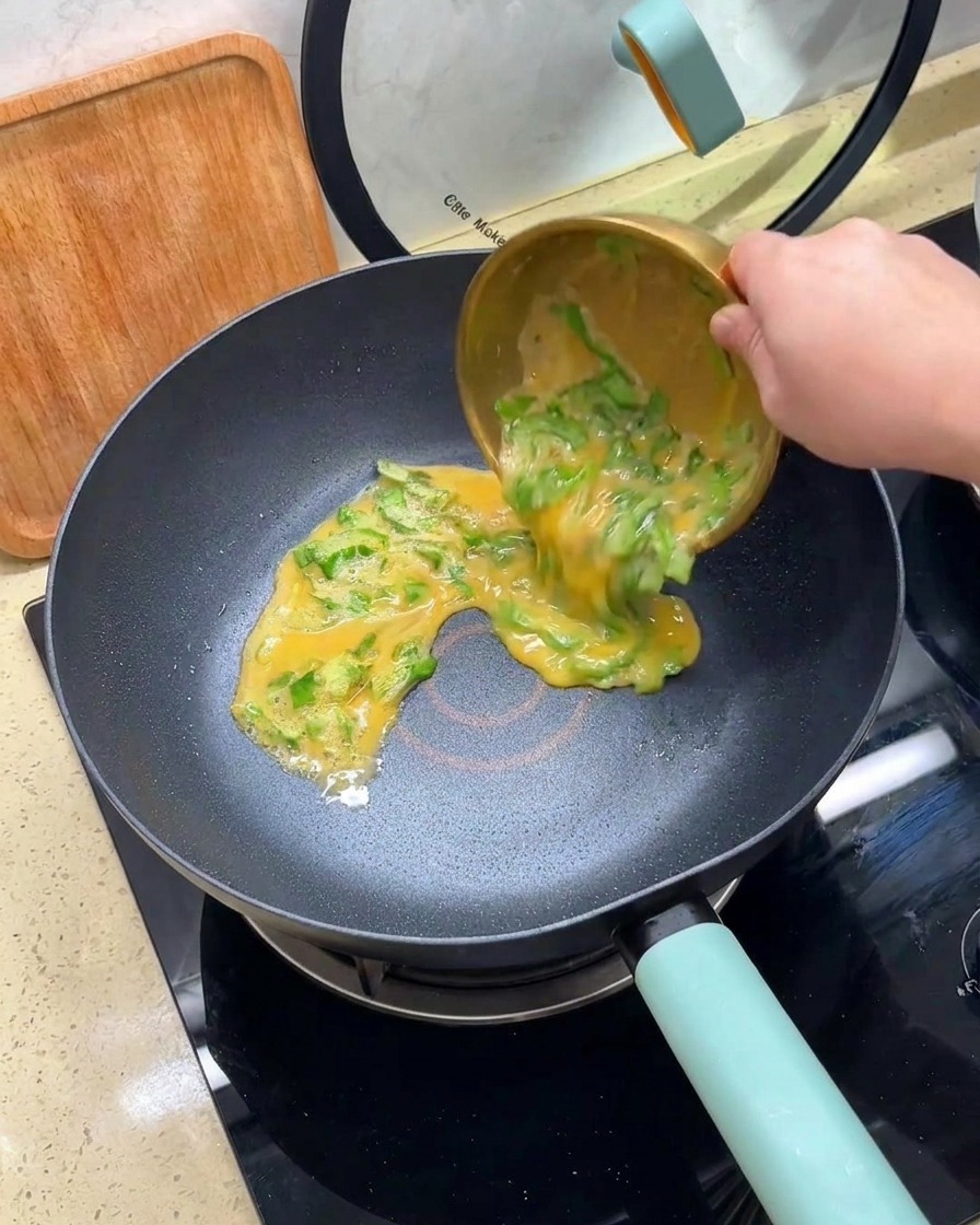 Eingießen einer Mischung aus verquirlten Eiern und gehacktem Salat aus einer goldenen Schüssel in eine heiße, beschichtete Pfanne.