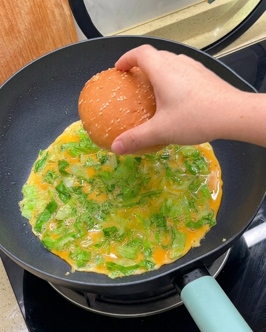 Eine Hand legt ein Sesabbrötchen mit der Schnittfläche nach unten auf die gärende Ei-Salat-Mischung in der Pfanne.