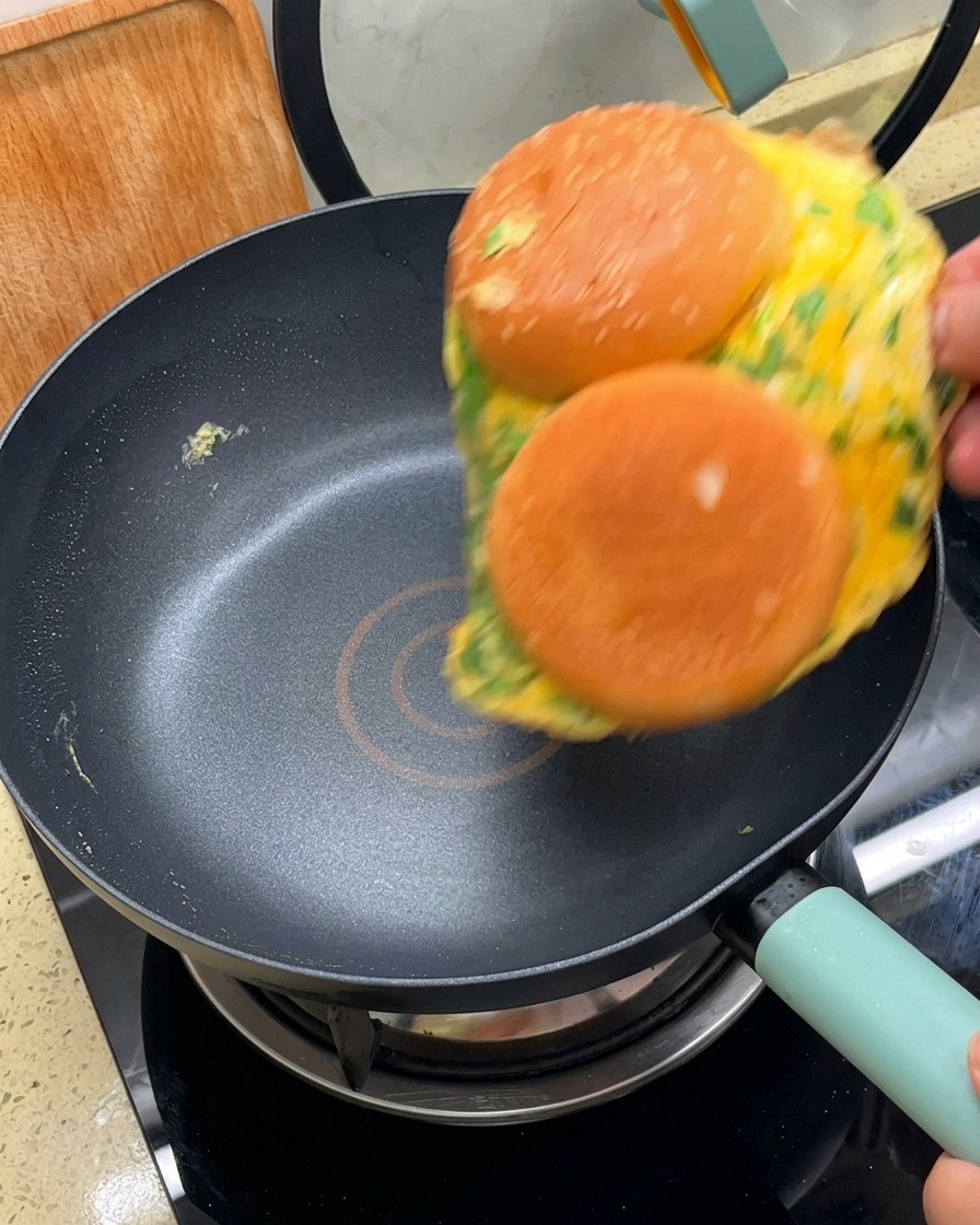 Ein Omelett mit zwei daran haftenden Burgerbrötchenhälften wird in einer schwarzen, beschichteten Pfanne gewendet.