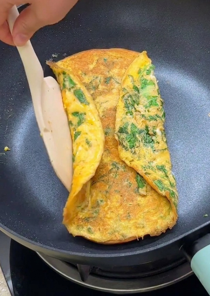Ein Pfannenwender klappt die Ränder eines grünen Salat-Omeletts über zwei getoastete Burgerbrötchenhälften in einer beschichteten Pfanne.