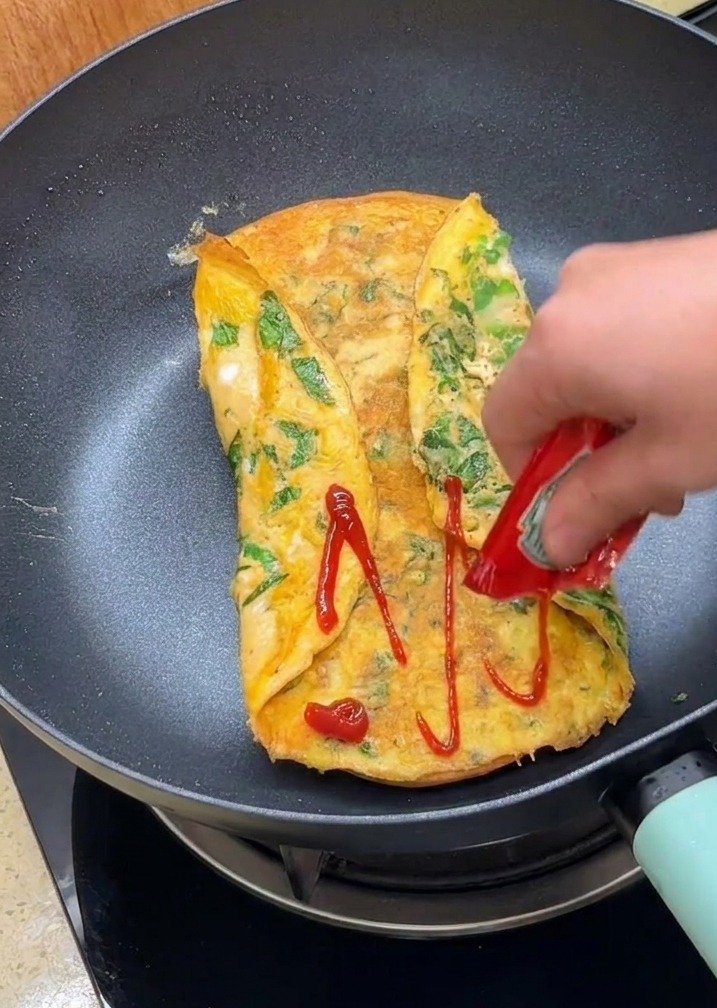 Eine Hand drückt roten Ketchup aus einem Päckchen auf die gefaltete Omelettoberfläche in der Pfanne.