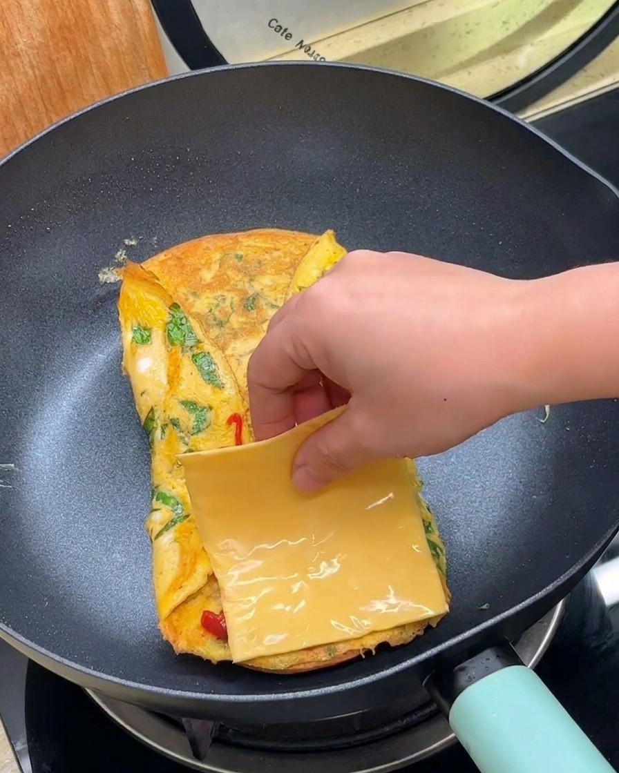Eine Hand legt eine quadratische Scheibe gelben Käse auf ein gefaltetes Ei in einer Bratpfanne.