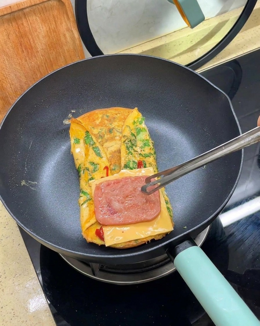 Eine Zange legt eine Scheibe gebratenes Fleisch über eine Scheibe Käse auf einem gefalteten Omelett in einer Pfanne.