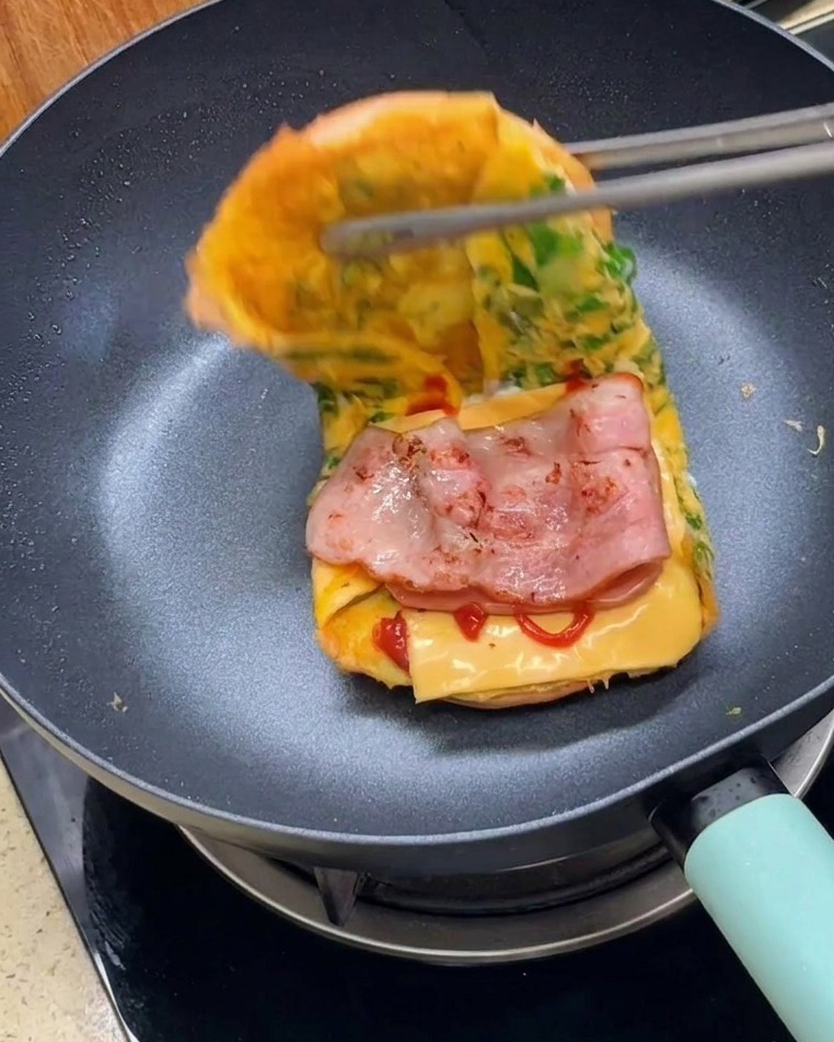 Eine Zange klappt die zwei Hälften des in Ei gehüllten Burgerbrötchens zusammen, um ein fertiges Sandwich zu bilden.