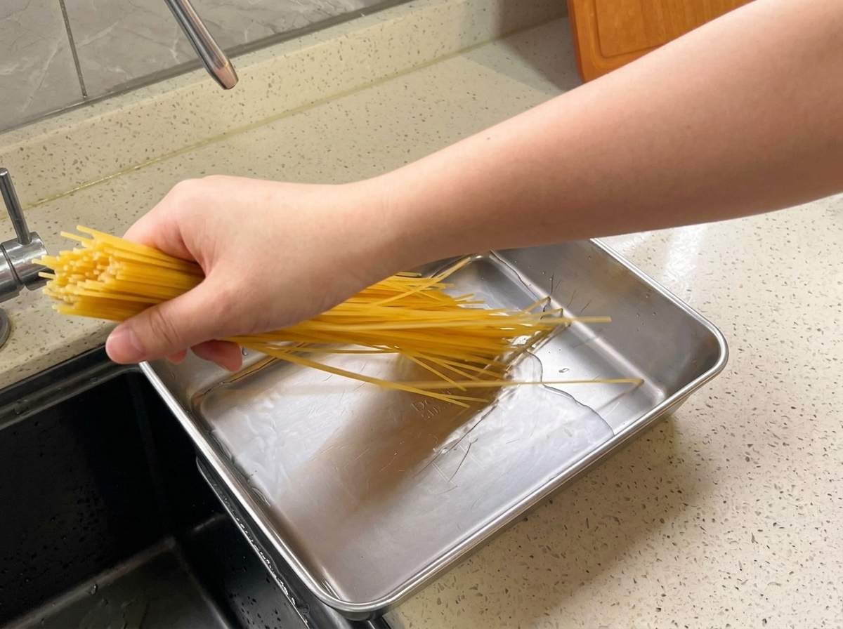 Eine Hand legt ein Bündel gelbe Spaghetti in eine flache Metallschale mit Wasser auf einer Küchenarbeitsplatte.