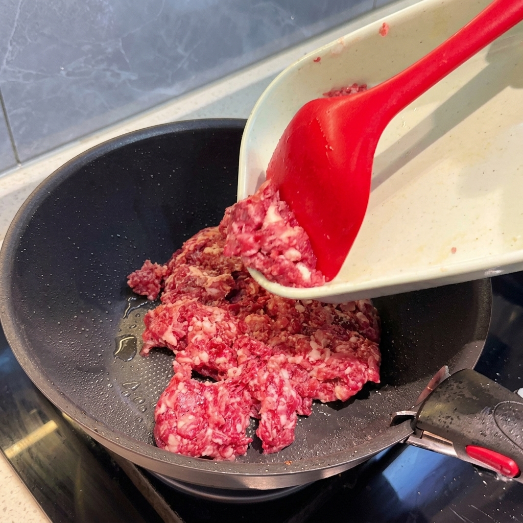 Rohes, grob gehacktes Mischfleisch wird mit einem roten Pfannenwender vom Schneidebrett in eine dunkle Pfanne geschoben.