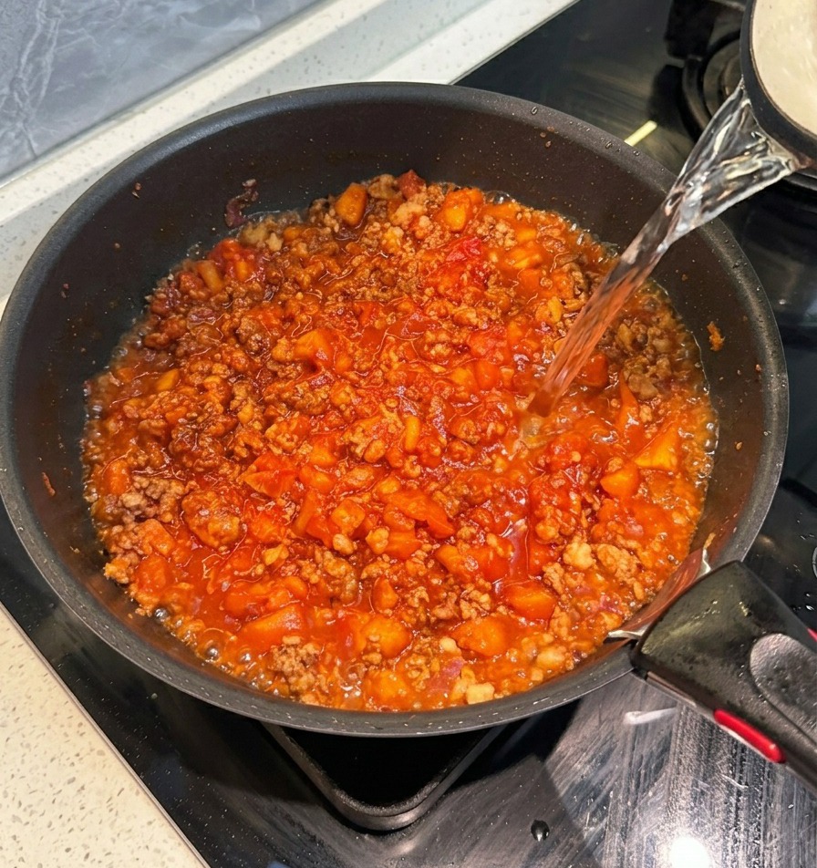 Klares heißes Wasser wird aus einem Wasserkocher in eine Pfanne mit dicker roter Tomaten-Fleischsauce gegossen.