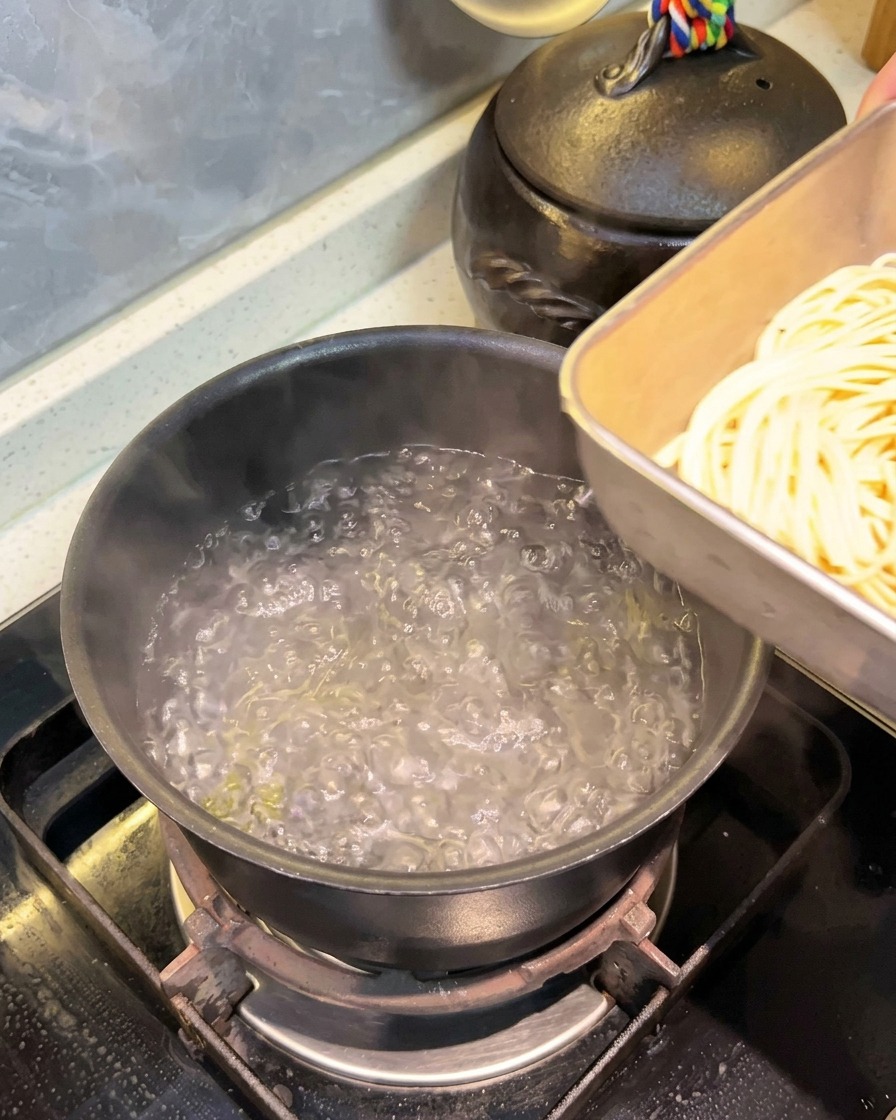 Eine Portion eingeweichte Spaghetti wird aus einer Metallschale in einen Topf mit kochendem Wasser gegeben.