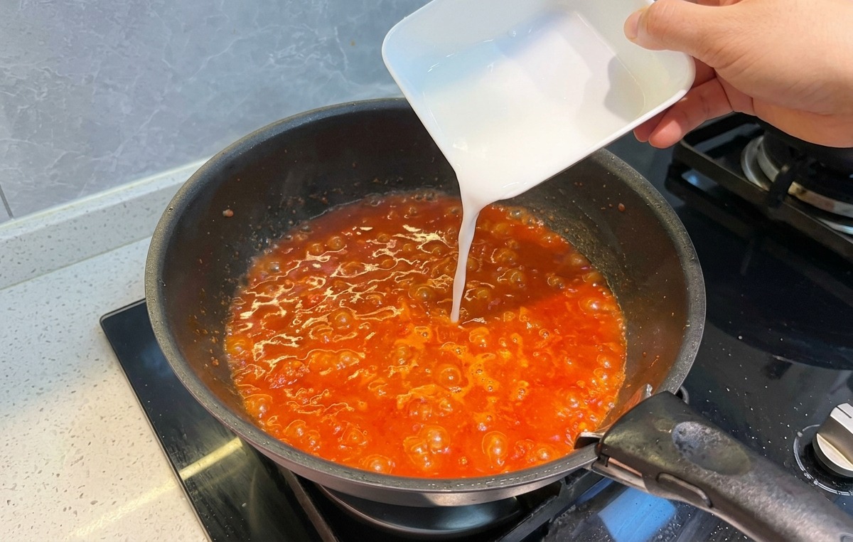 Eine milchig-weiße Stärkemischung wird aus einer kleinen Schale in die blubbernde Tomaten-Fleischsauce gegossen.