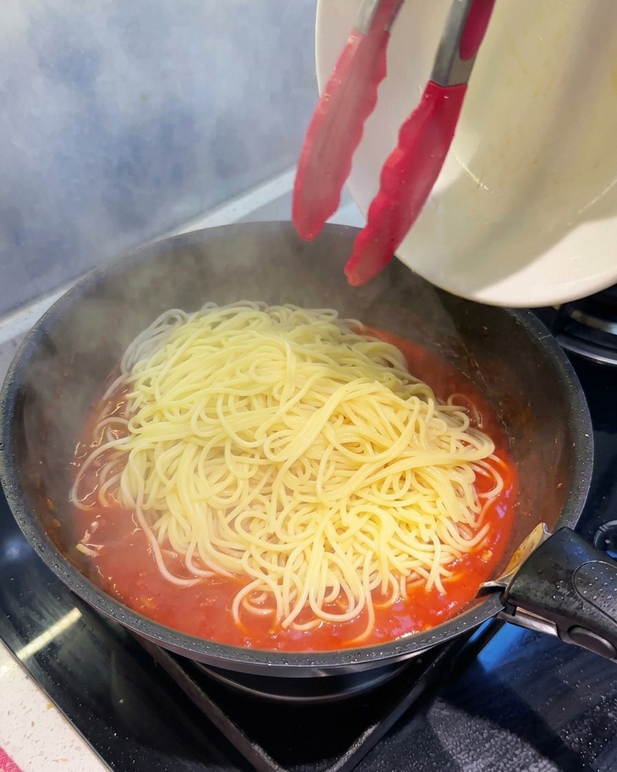 Mit einer Küchenzange werden dampfende, frisch gekochte Spaghetti in die Pfanne mit der dicken Fleischsauce gegeben.