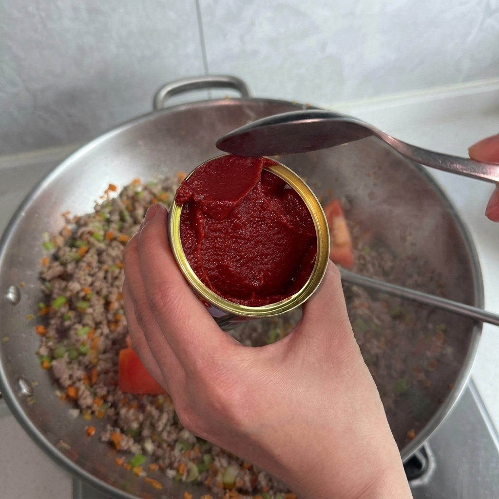 Rotes Tomatenmark wird mit einem Löffel in den Wok zu Fleisch und Gemüse gegeben.