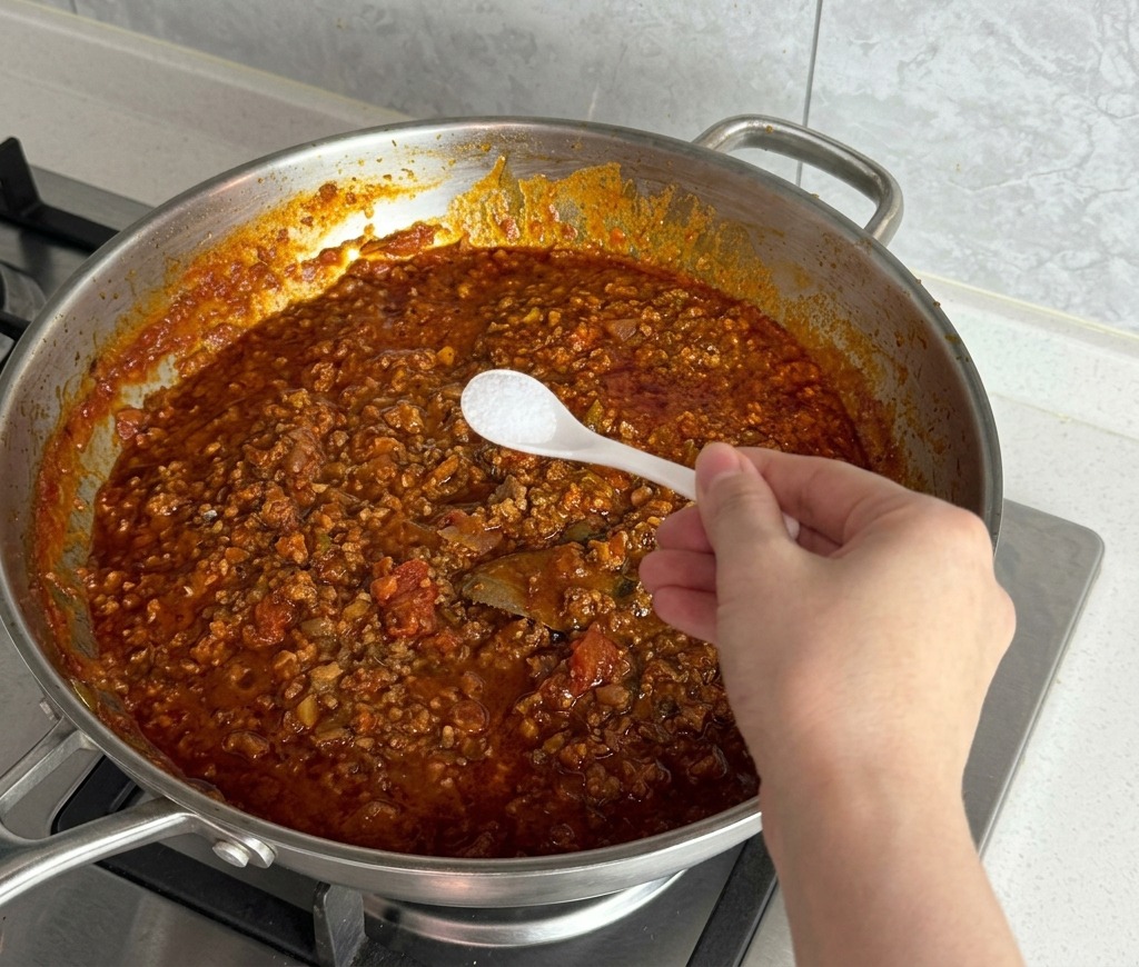 Salz wird mit einem kleinen Löffel zur köchelnden Fleischsauce gegeben.