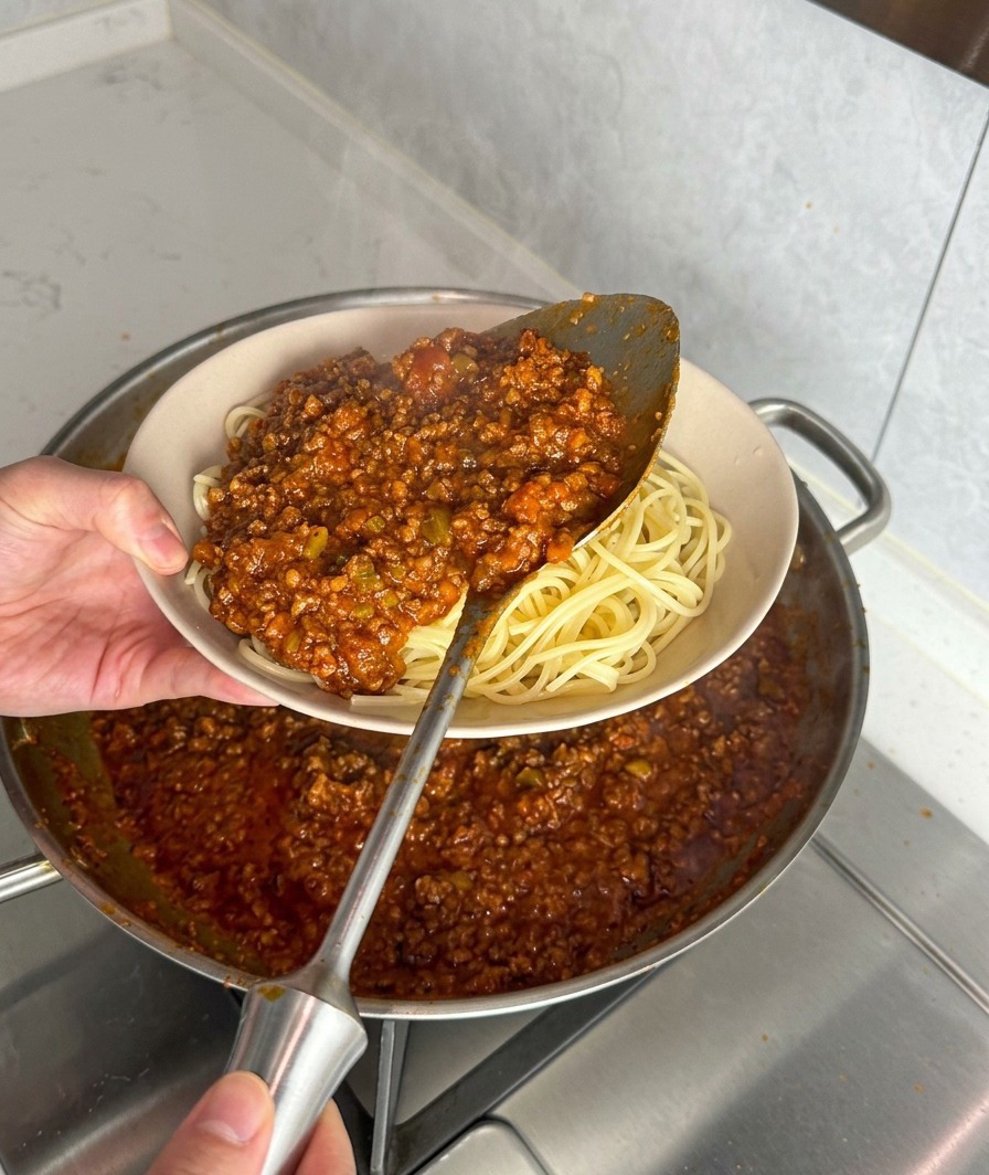 Eine Kelle Fleischsauce wird über frisch gekochten Spaghetti serviert.