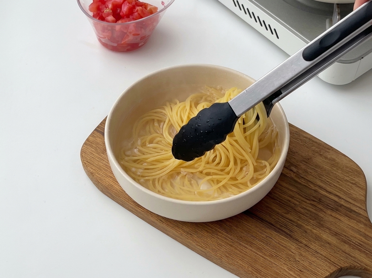 Eine schwarze Küchenzange hebt gekochte Spaghetti aus dem heißen Wasser in eine Schüssel mit kaltem Wasser.