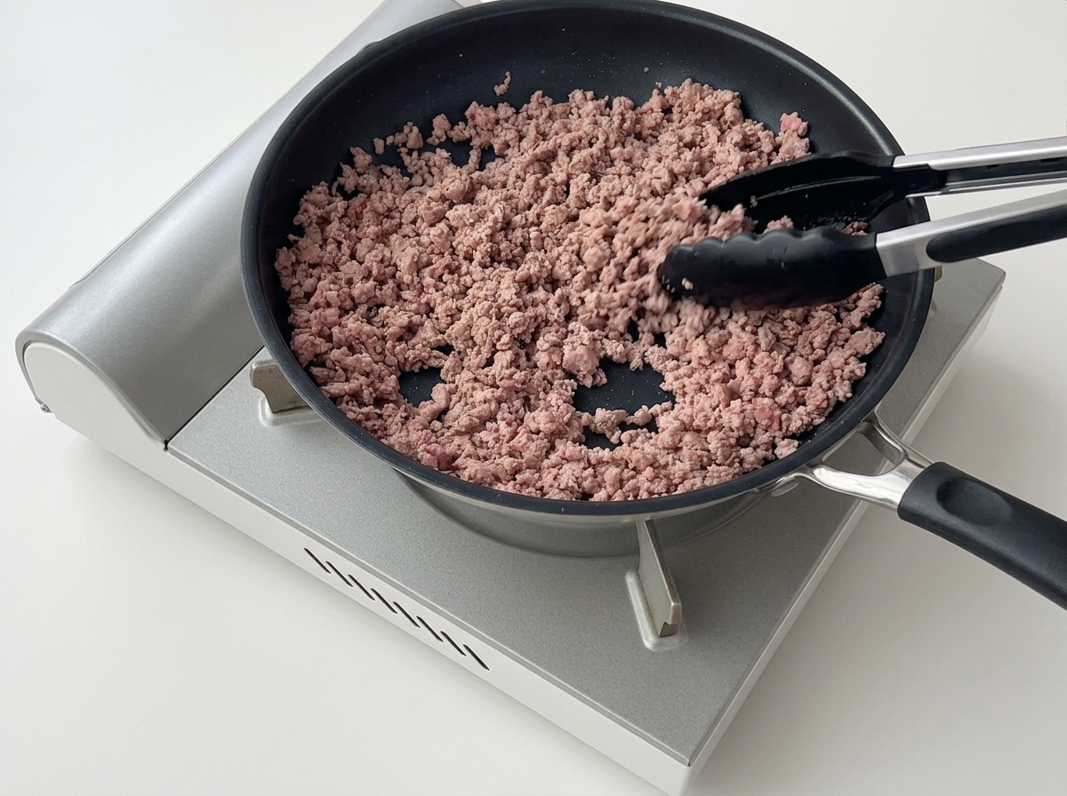 Küchenzange zerkleinert braun gebratenes Hackfleisch in der Pfanne.
