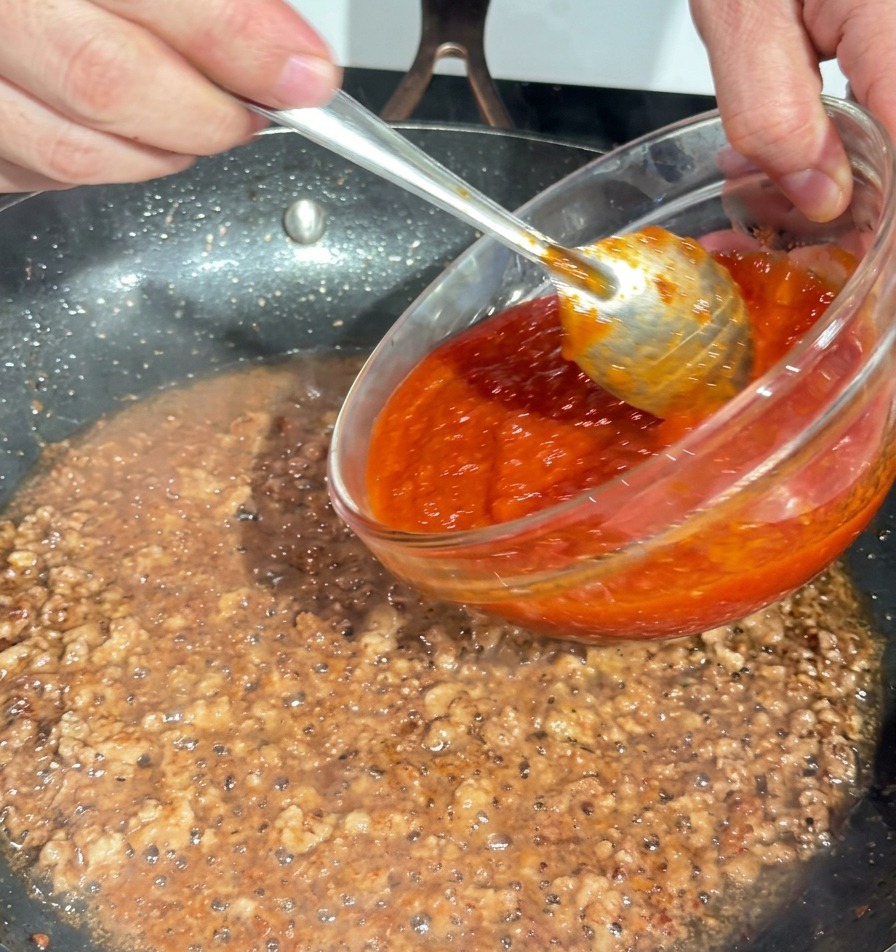 Dicke rote Tomatensauce wird aus einer Glasschüssel in die Pfanne zum Fleisch gegossen.