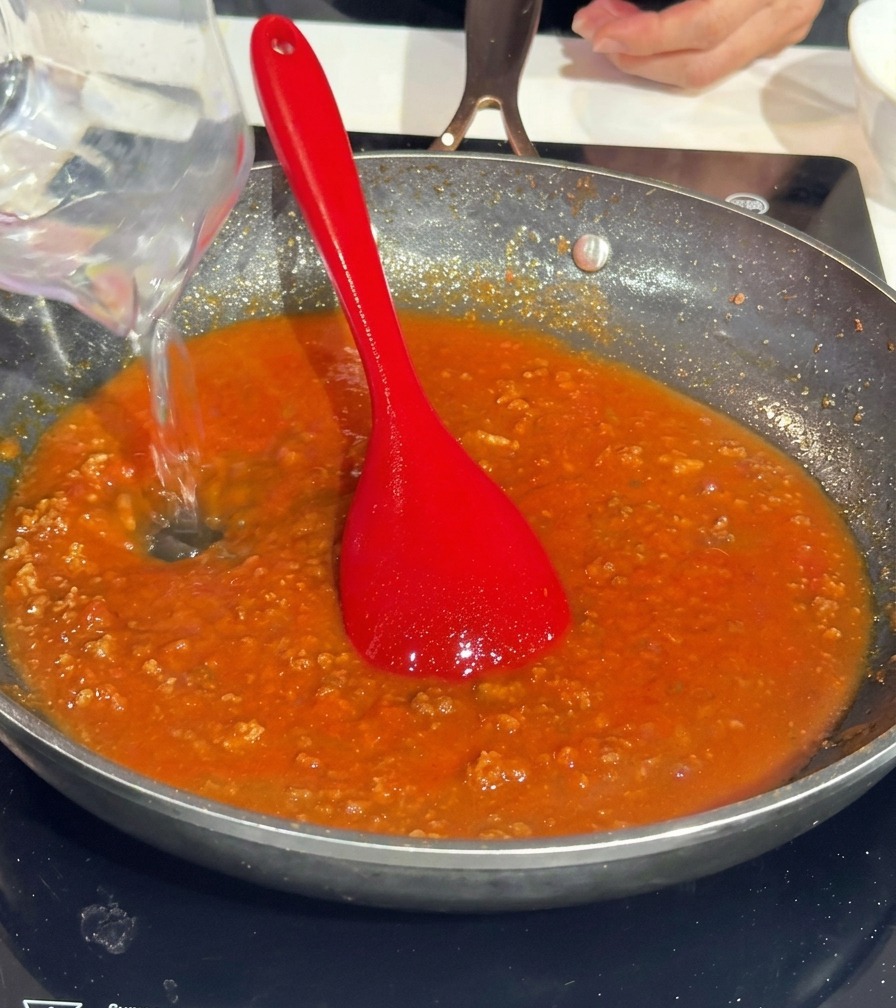 Wasser wird aus einem Messbecher in die Pfanne mit der Tomaten-Fleischsauce gegossen.