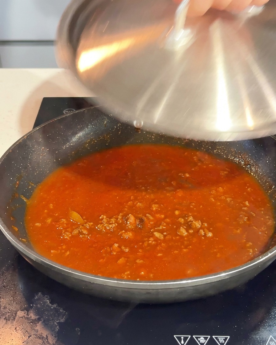 Eine Hand legt einen Edelstahldeckel auf die Pfanne mit der blubbernden Bolognese.