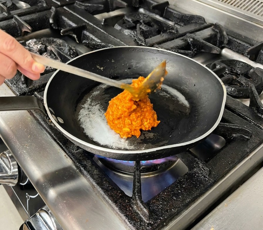 Ein großer Löffel rötliche Fleischsauce wird in eine erhitzte Pfanne gegeben.