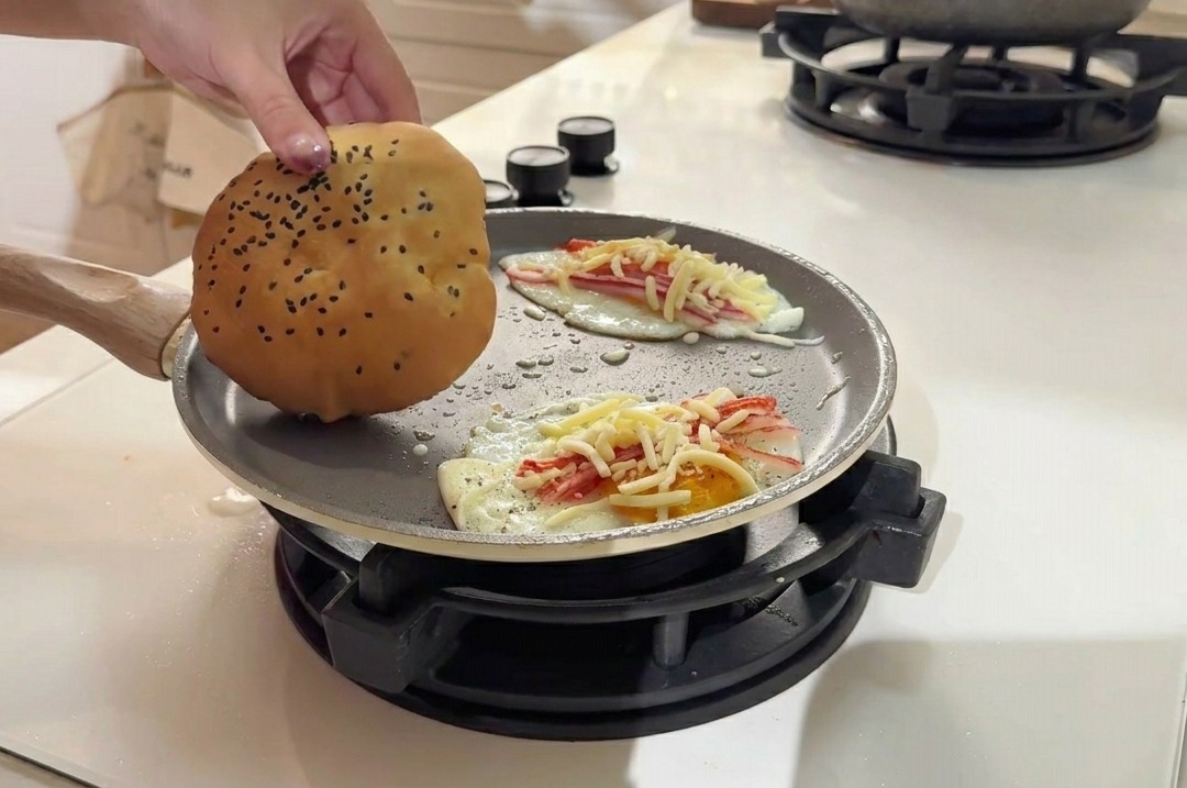 Ein goldenes Sesam-Burgerbrötchen wird neben den Eier-Patties auf die Pfannenoberfläche gelegt.
