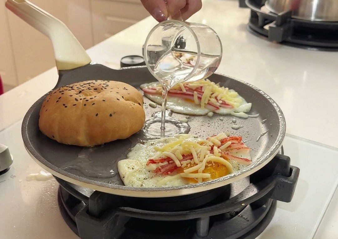 Klares Wasser wird aus einem kleinen Glas in die heiße Pfanne zwischen das Brötchen und die Eier gegossen.