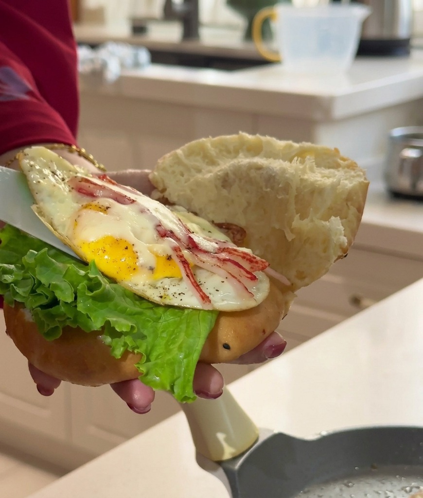 Eine Hand hält ein offenes gedämpftes Brötchen mit Salat, während ein Pfannenwender das Ei-Patty hineinschiebt.