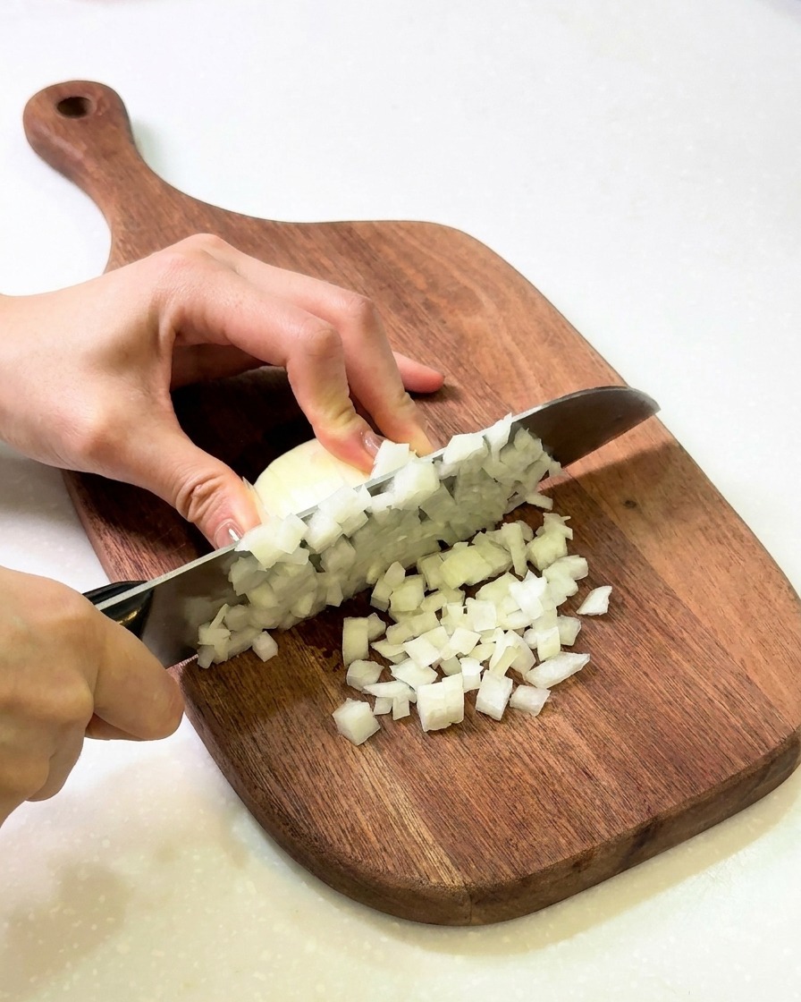 Hände schneiden eine weiße Zwiebel in feine Würfel auf einem Holzschneidebrett.