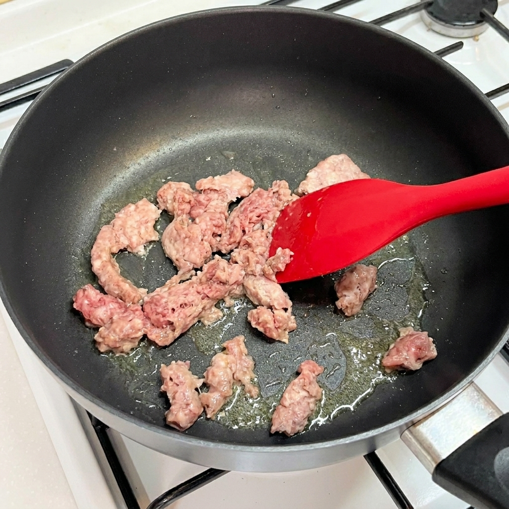 Rohes rosa Rinderhackfleisch wird in einer heißen Pfanne mit einem roten Silikonspatel zerkleinert.