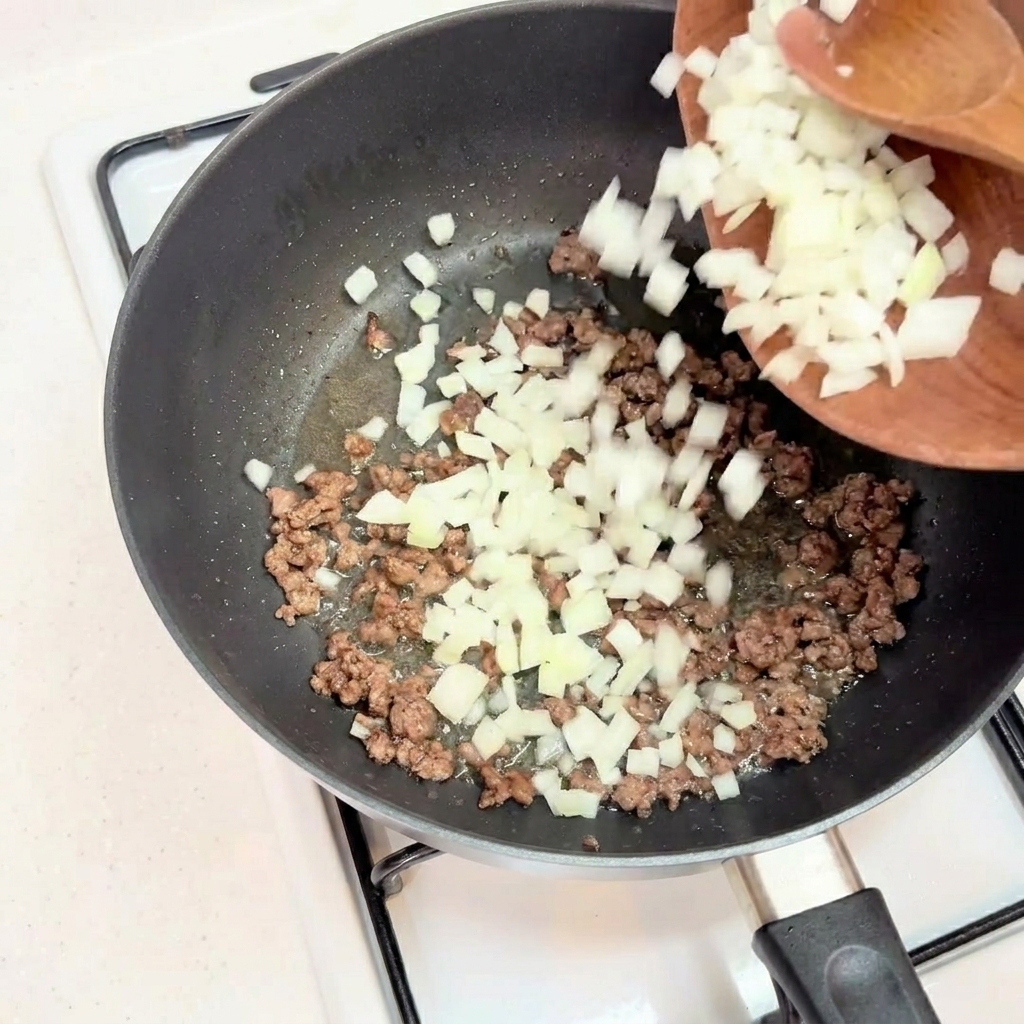 Gehackte weiße Zwiebeln werden zu gebräuntem Rinderhack in einer schwarzen Pfanne gegeben.