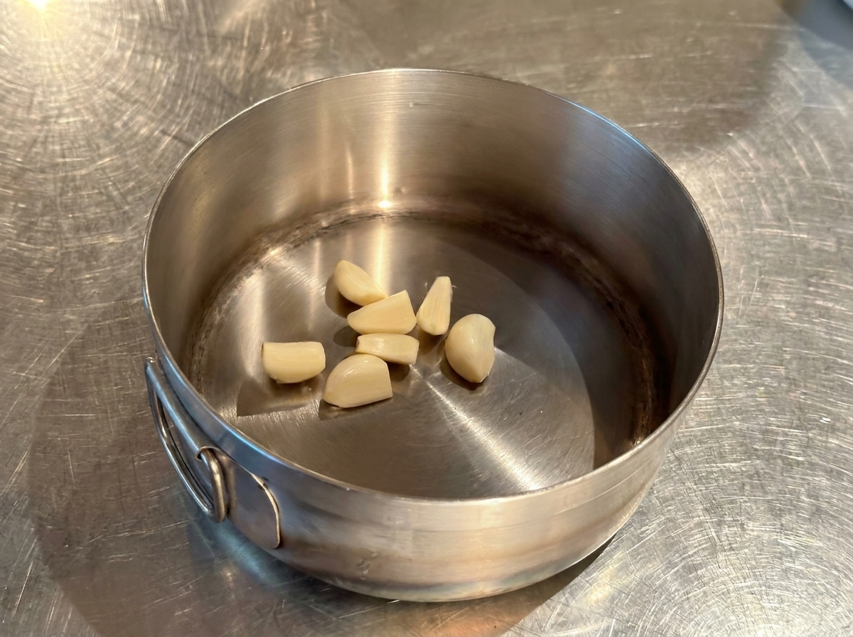 Mehrere ganze geschälte Knoblauchzehen liegen am Boden eines kleinen, trockenen Edelstahltopfes.