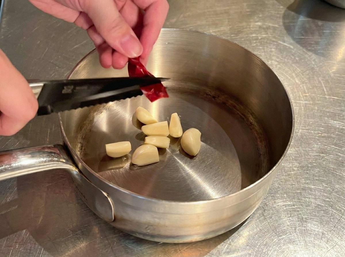 Hände schneiden eine getrocknete rote Chilischote mit einer Küchenschere in Stücke über einem Topf mit Knoblauch.
