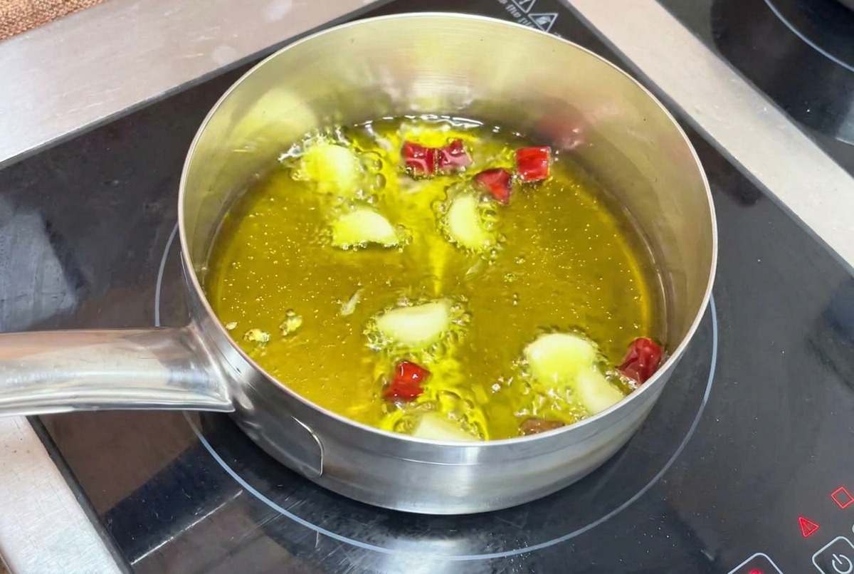 Knoblauchzehen und gehackte getrocknete Chilis brutzeln sanft in heißem Olivenöl in einem kleinen Metalltopf auf einem Induktionsherd.
