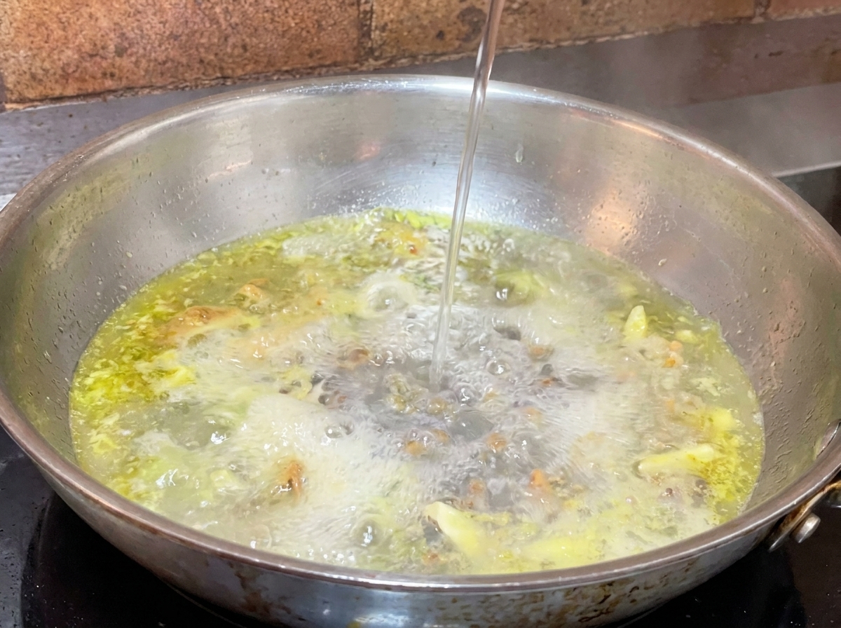 Ein dünner Strahl Nudelwasser wird in eine heiße Pfanne mit brodelndem Öl und Knoblauch gegossen.