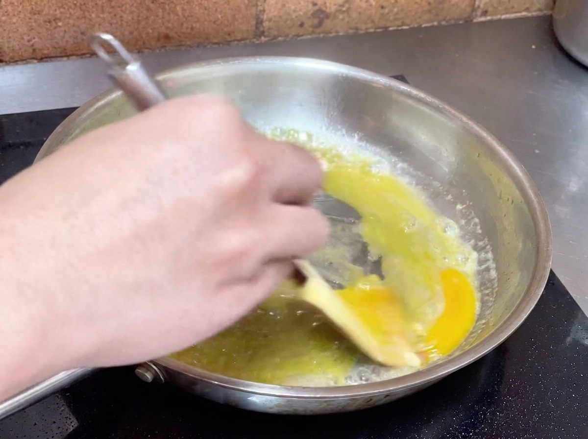 Eine Hand rührt eine gelbe Öl-Wasser-Mischung kräftig mit einem Spatel in einer Pfanne um, um eine cremige Sauce zu erzeugen.
