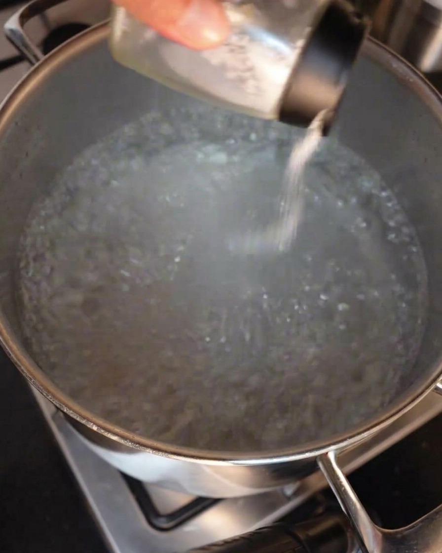 Eine Hand streut Salz aus einem Streuer in einen großen Edelstahltopf mit sprudelnd kochendem Wasser.