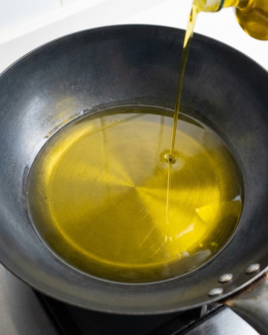 Ein goldener Strahl Olivenöl wird aus einer Flasche in die Mitte einer dunklen Pfanne gegossen.