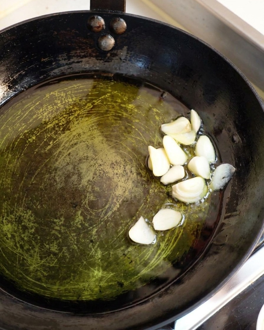 Dick geschnittene Knoblauchscheiben brutzeln sanft in Olivenöl in einer dunklen Pfanne.