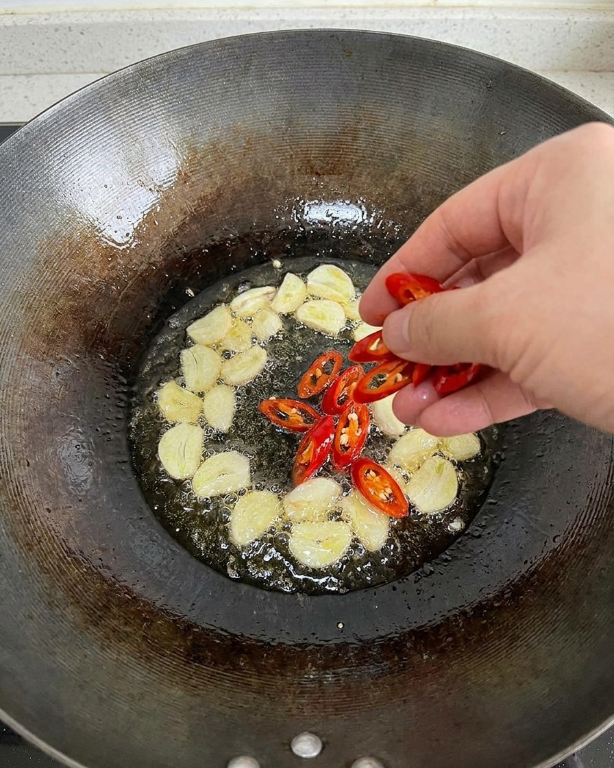 Eine Hand gibt frisch gehackte rote Chilischoten in eine Pfanne mit brutzelndem Knoblauch.