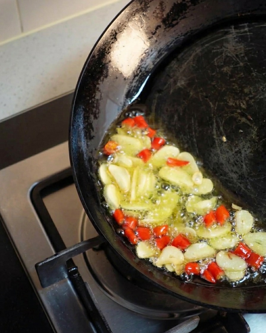 Ein schräg gehaltener Wok, in dem Knoblauch und Chili in einer Pfütze aus goldenem Öl sprudelnd frittiert werden.