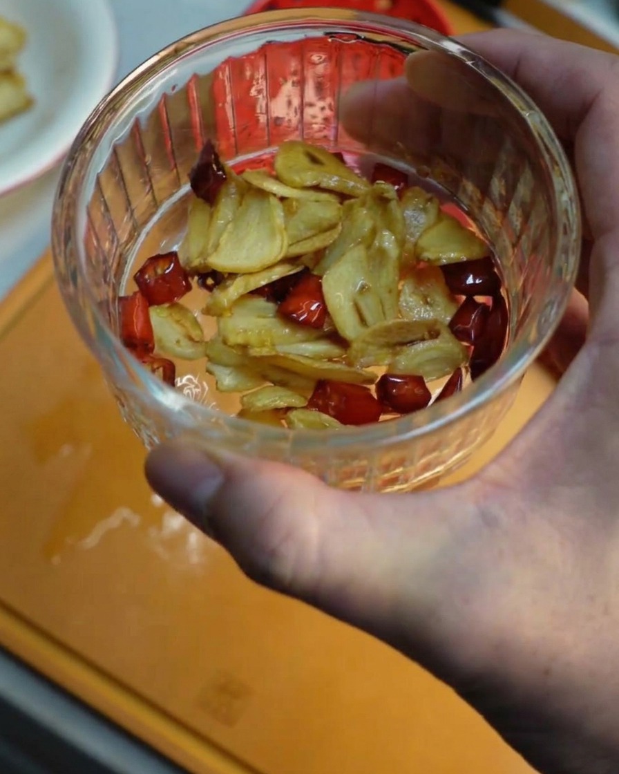 Eine Hand hält eine kleine Glasschüssel mit knusprig frittiertem Knoblauch und Chilistücken.