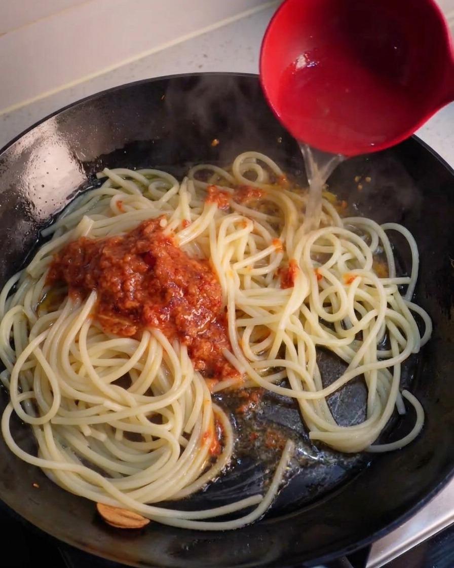 Eine rote Schöpfkelle gießt Nudelwasser in eine Pfanne über Spaghetti und einen Klecks Sauce.