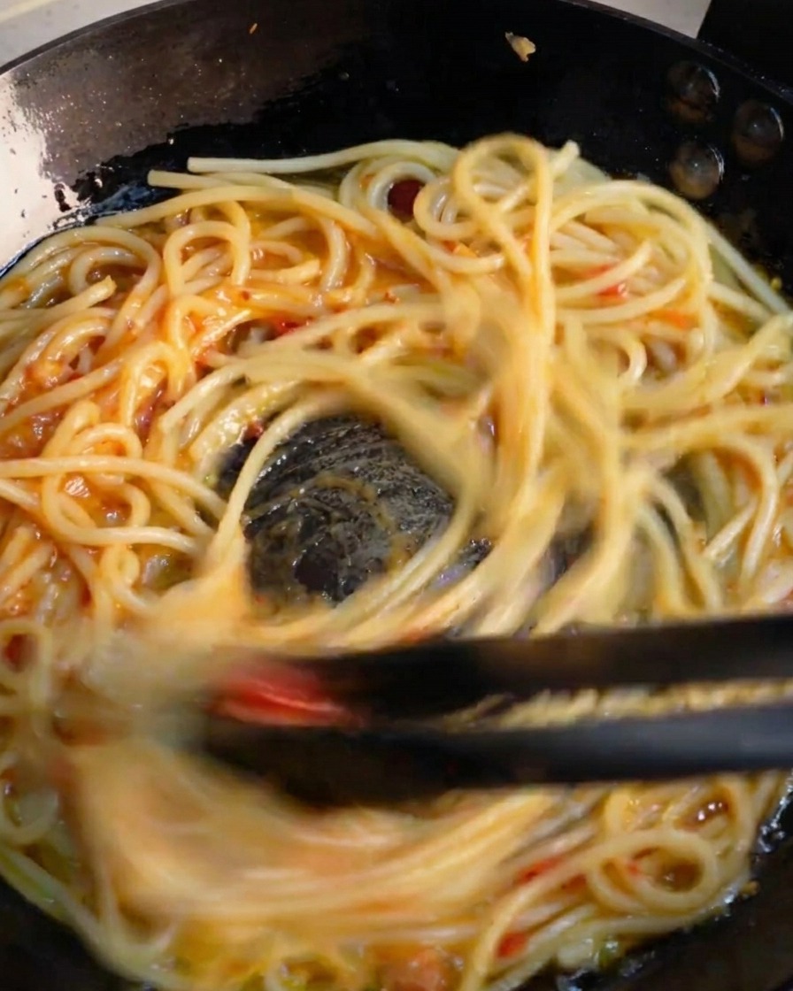 Schwarze Küchenzangen schwenken Spaghetti kräftig in der Pfanne, um die Sauce zu emulgieren.